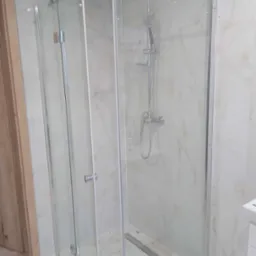 Kabina prysznicowa z przezroczystego szkła z chromowanymi okuciami, widoczny odpływ liniowy w posadzce, marmurowe płytki na ścianach i podłodze, drzwi wejściowe w tle.