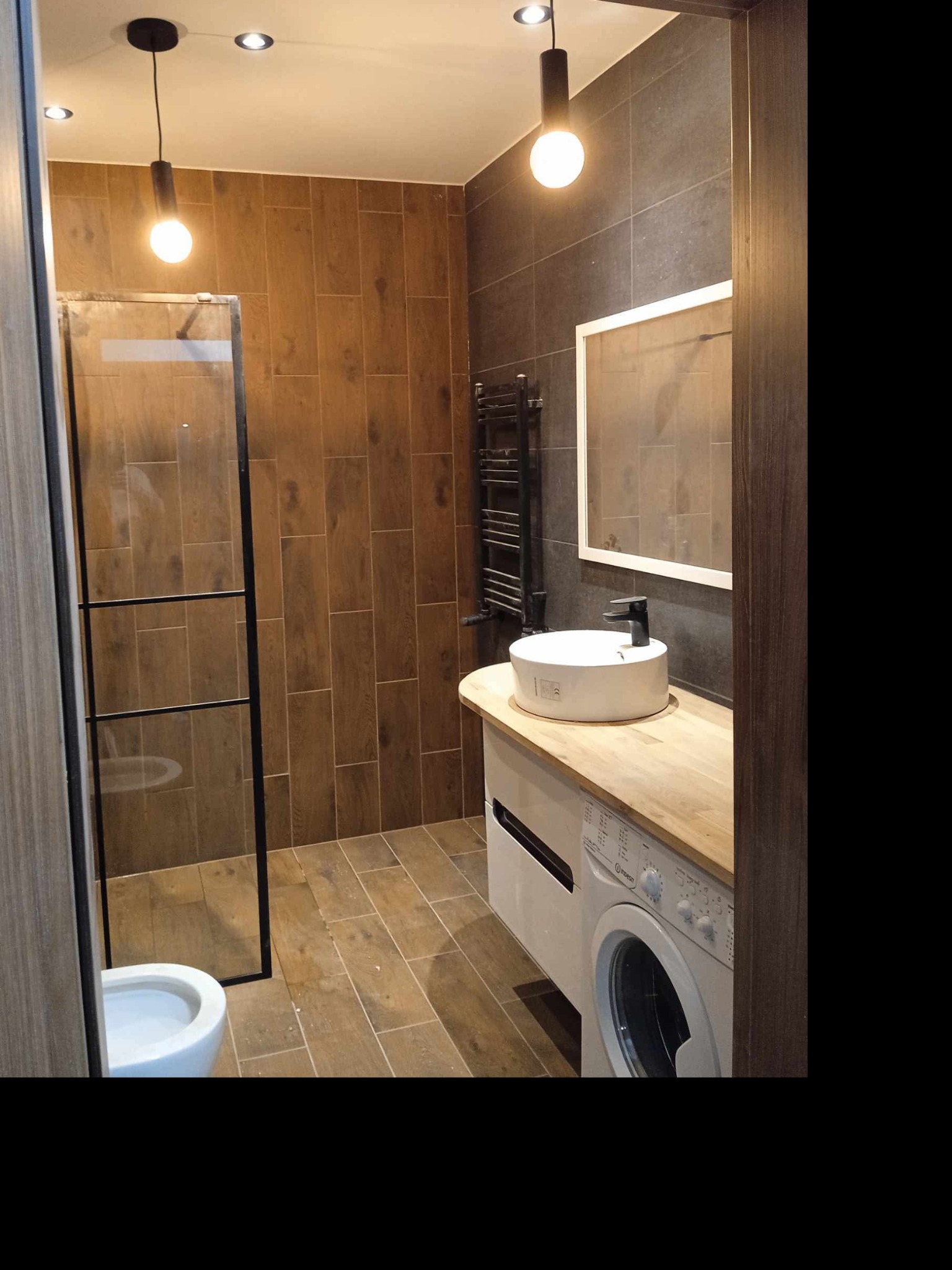 Nowoczesna łazienka z kabiną prysznicową typu walk-in, umywalką nablatową na blacie z pralką pod spodem, brązowe płytki imitujące drewno na ścianach i podłodze, oraz szare płytki na jednej ze ścian...