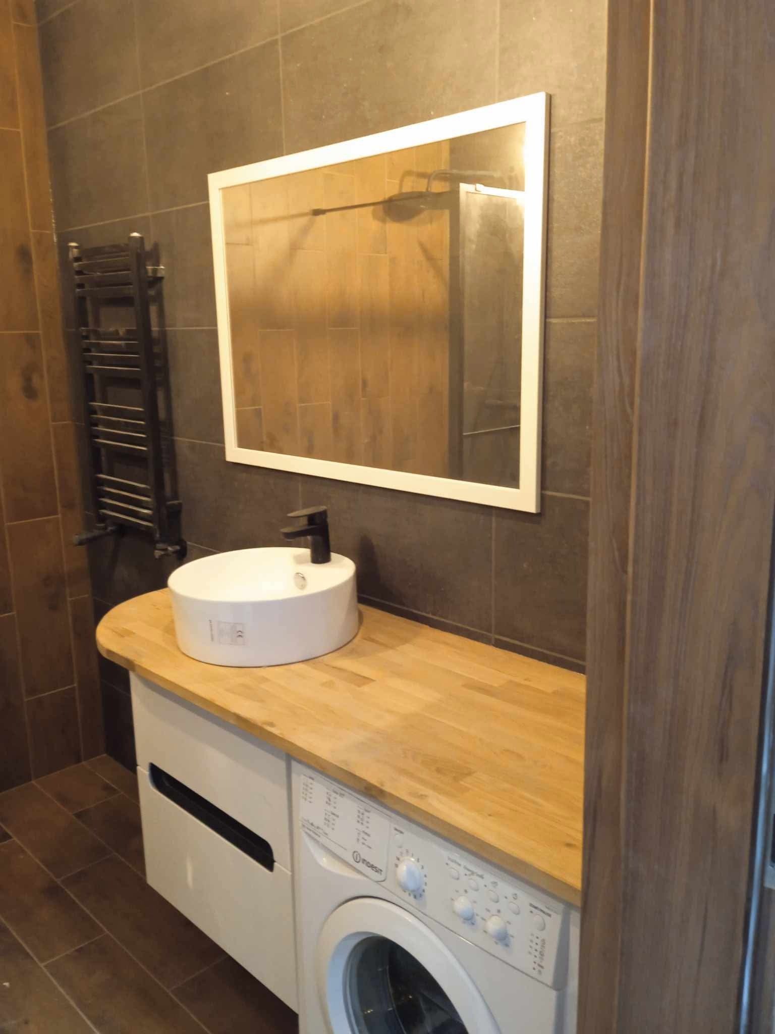 Nowoczesna łazienka z umywalką nablatową na drewnianym blacie, pod którym znajduje się pralka, lustro w białej ramie na tle szarych płytek i ciemny grzejnik drabinkowy.