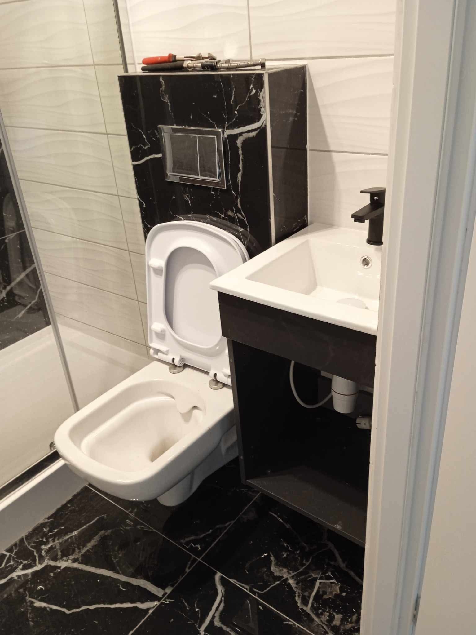 Nowoczesna łazienka z czarno-białymi płytkami imitującymi marmur, wisząca toaleta z zabudowaną spłuczką i umywalką z czarną armaturą.