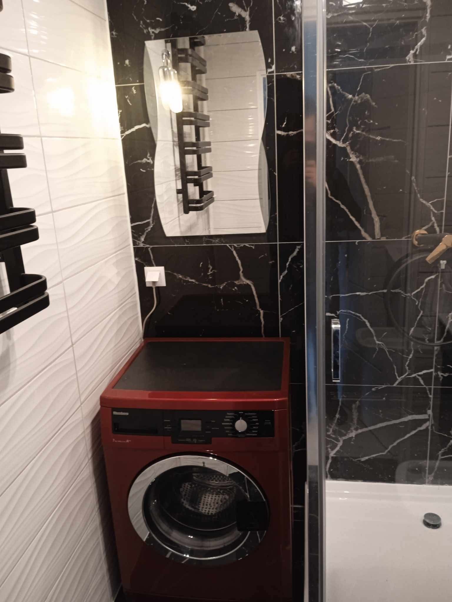 Nowoczesna łazienka z czerwonym automatem do prania, biało-czarne płytki imitujące marmur, lustro z oświetleniem i kabina prysznicowa.
