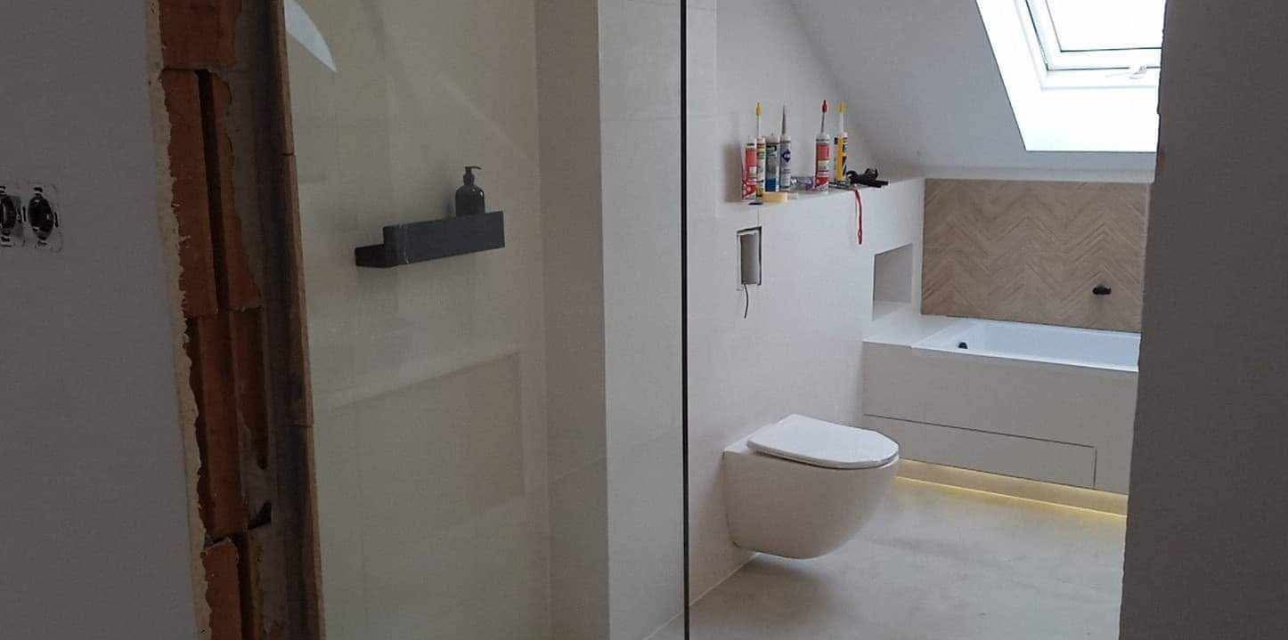 Nowoczesna łazienka w trakcie wykończenia: wisząca toaleta, wanna obudowana płytkami, drewniana mozaika na ścianie, widoczne materiały budowlane i narzędzia.