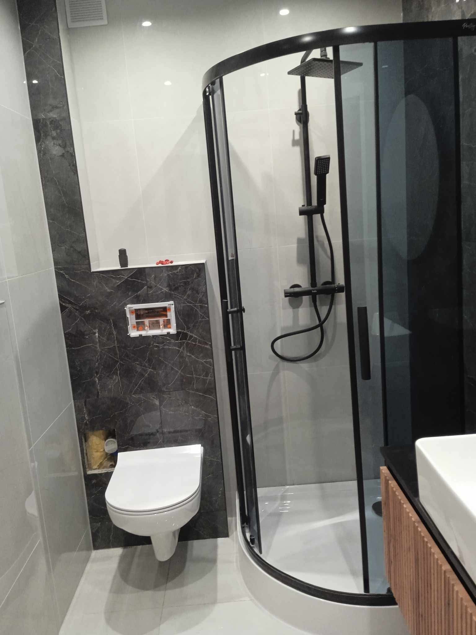 Nowoczesna łazienka z kabiną prysznicową typu walk-in z czarnymi profilami, podwieszaną toaletą na tle płytek imitujących marmur i umywalką z drewnianą szafką.