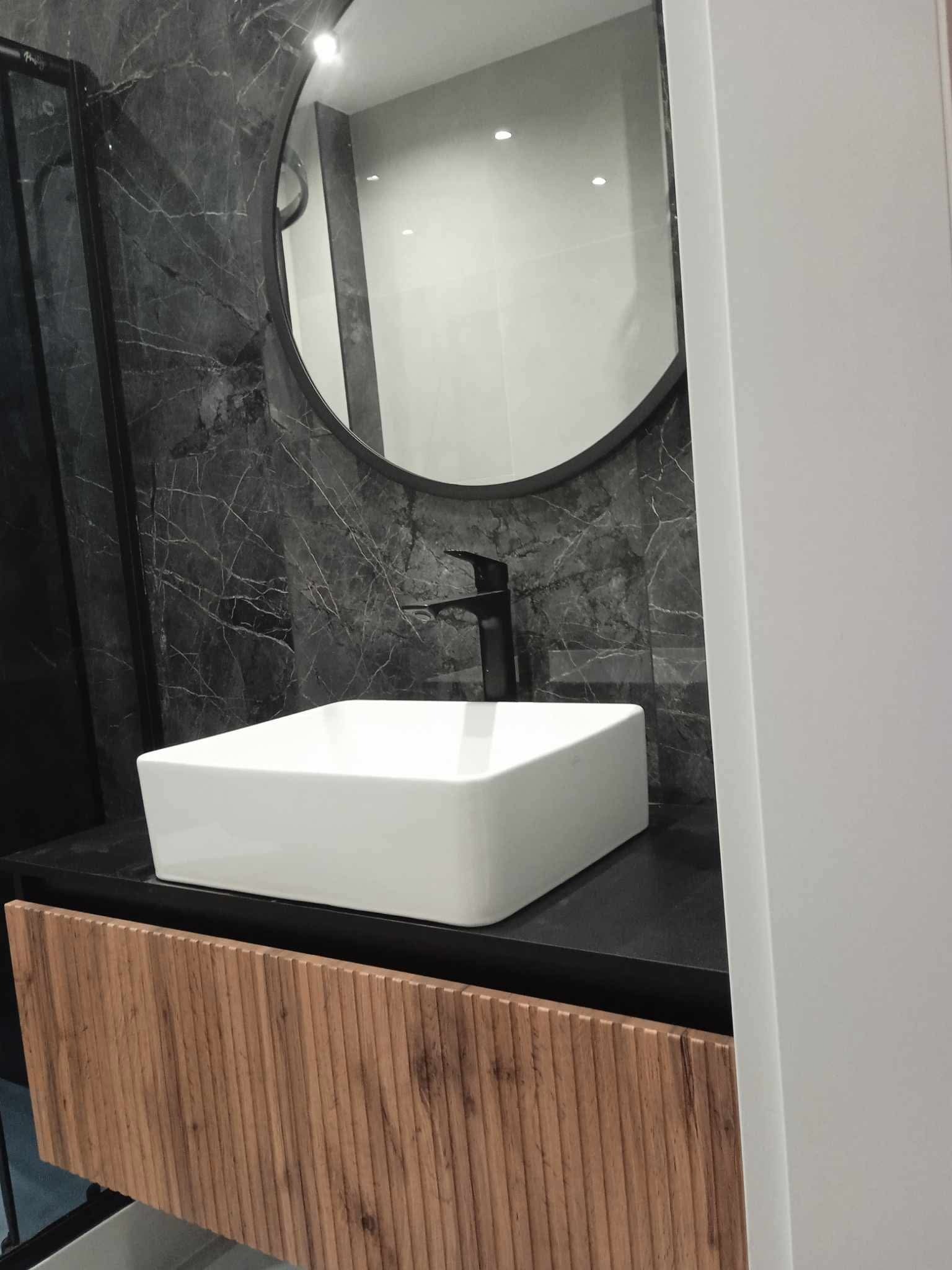 Nowoczesna łazienka z umywalką nablatową o kwadratowym kształcie, czarną armaturą i okrągłym lustrem na tle ściany z szarego marmuru z wyraźnym użyleniem.