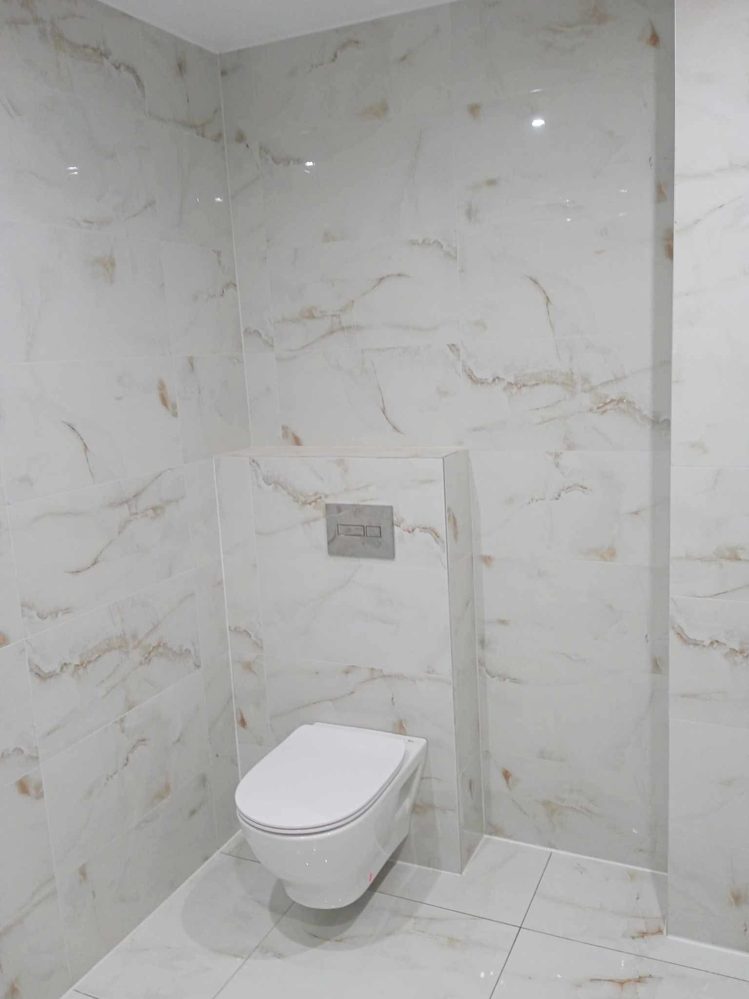 Wykończona łazienka z białymi płytkami imitującymi marmur, wisząca toaleta z ukrytym stelażem podtynkowym, minimalistyczny styl.