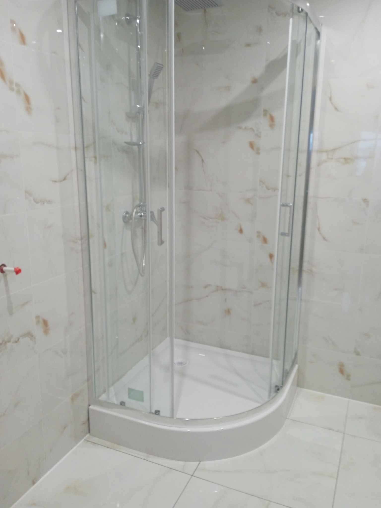 Narożna kabina prysznicowa z przezroczystego szkła z chromowaną armaturą, zamontowana w łazience wyłożonej białymi płytkami imitującymi marmur z delikatnym, pomarańczowym żyłowaniem.