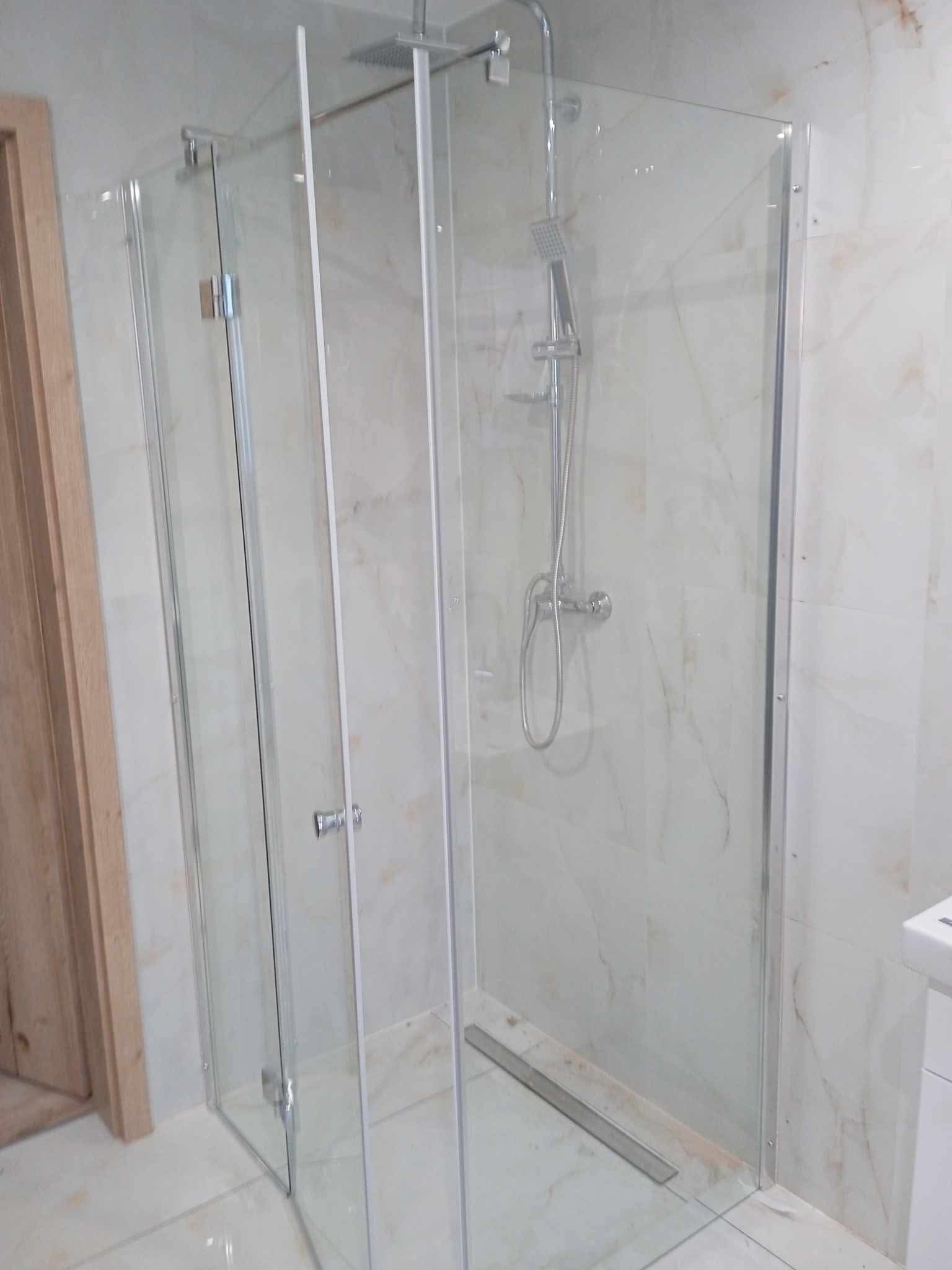 Kabina prysznicowa z przezroczystego szkła z chromowanymi okuciami, widoczny odpływ liniowy w posadzce, marmurowe płytki na ścianach i podłodze, drzwi wejściowe w tle.