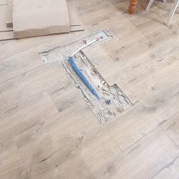 Dmytro kokoshko - Prace hydrauliczne w Wałbrzychu: Wykute miejsce w podłodze pod rury, widoczne fragmenty instalacji. Podłoga imitująca drewno, w tle meble.