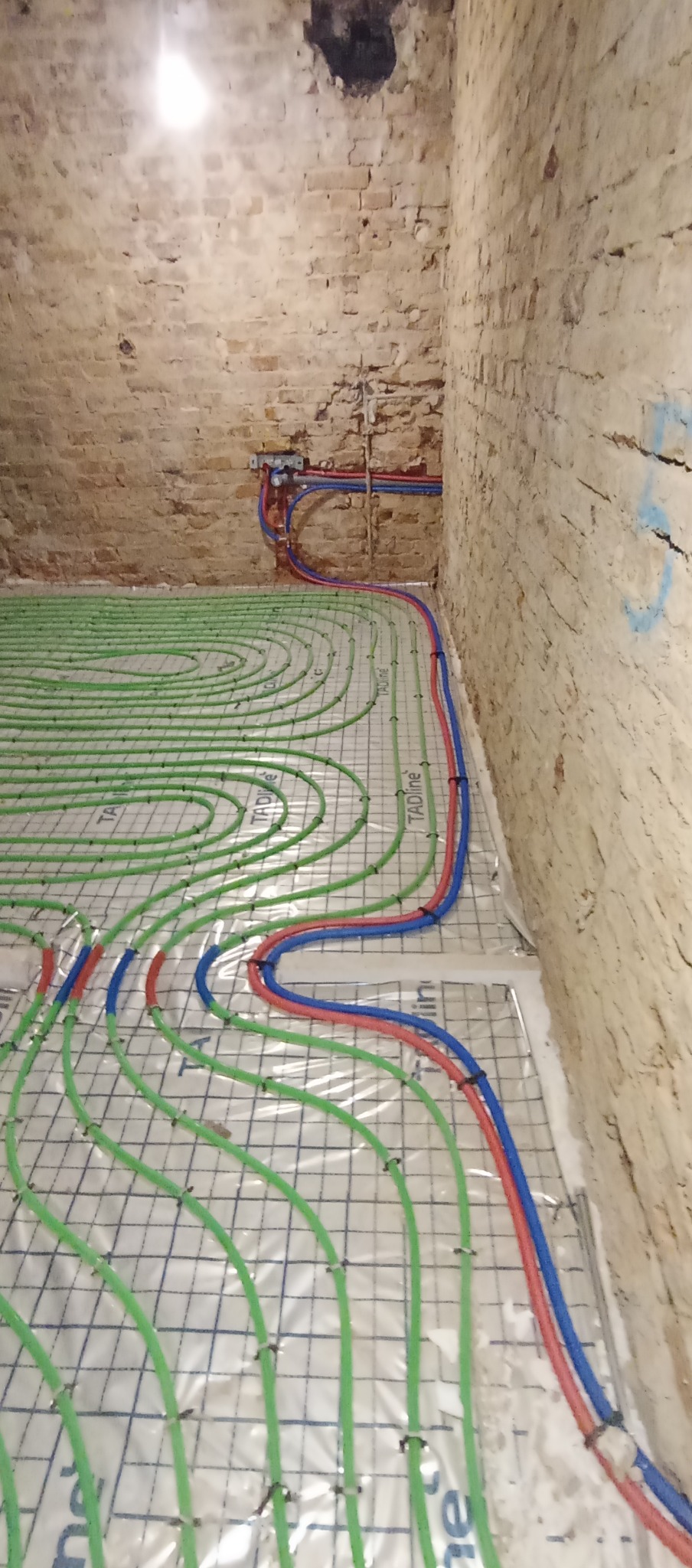 Ułożone rury ogrzewania podłogowego w pomieszczeniu z ceglanymi ścianami. Widoczne zielone pętle rur na izolacji, oraz czerwone i niebieskie rury przy ścianie.