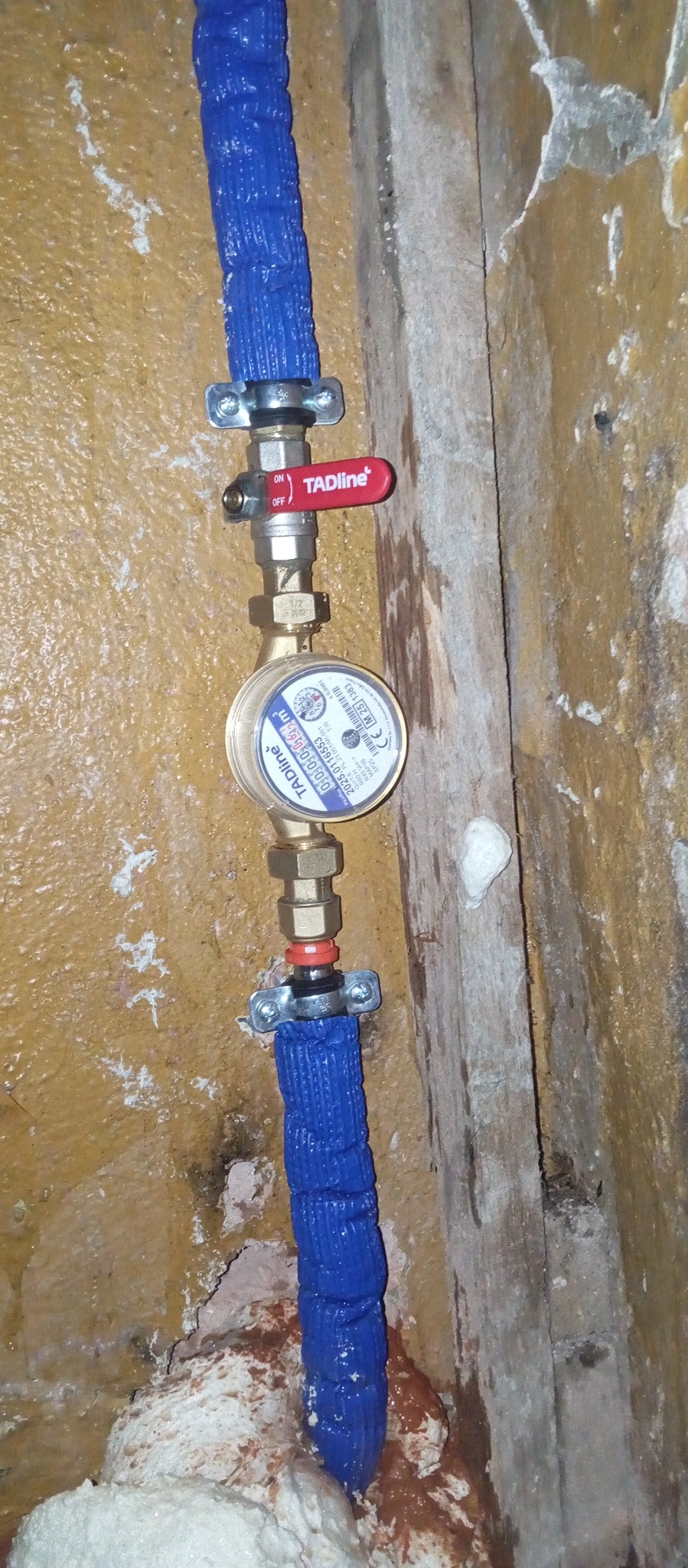 Zamknięty zawór z licznikiem wody Tadline, zamontowany na niebieskich rurach w pomieszczeniu o surowych ścianach. Widoczne mocowania i elementy instalacji.