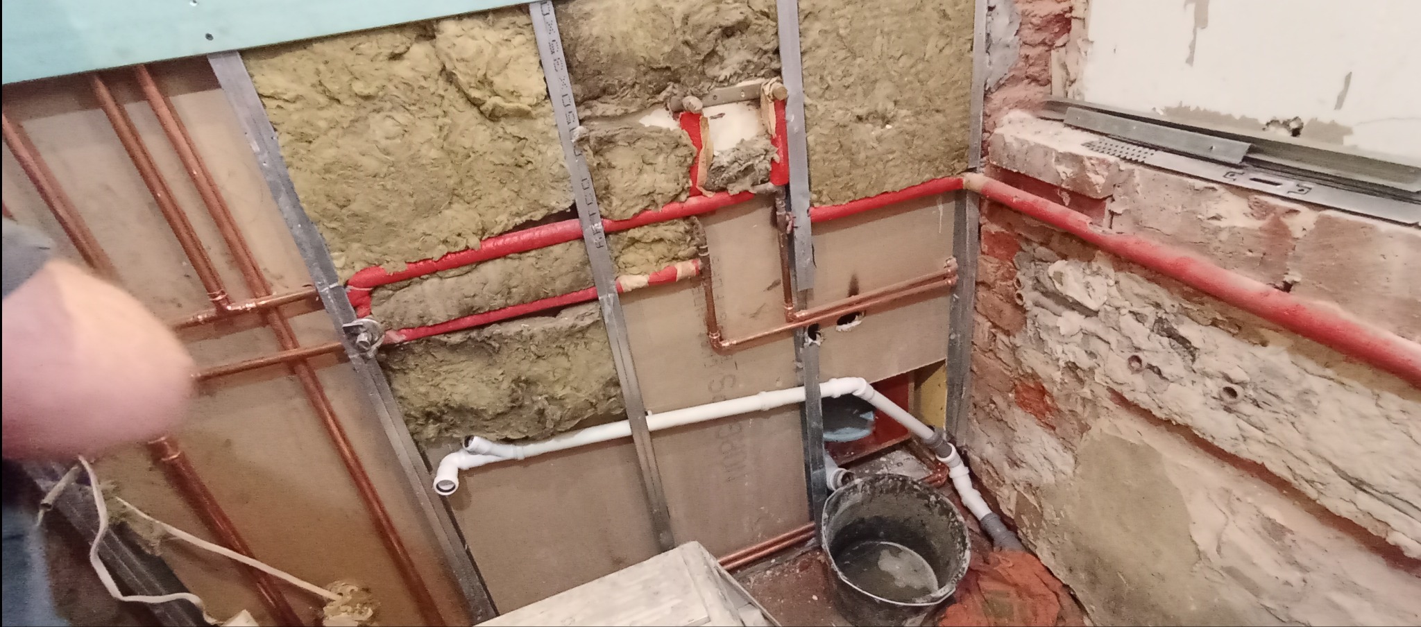 Instalacja wodna w trakcie remontu łazienki: miedziane rury, izolacja z wełny mineralnej, białe rury PCV, surowa ściana z cegły. Widoczny etap prac.