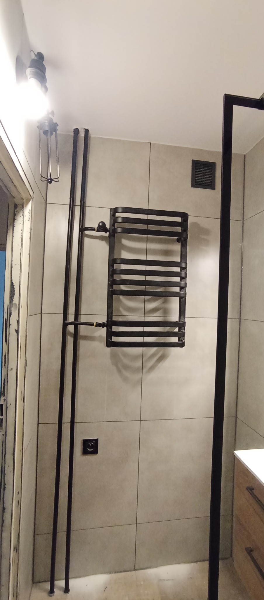 Czarny, drabinkowy grzejnik łazienkowy na tle beżowych płytek. Widoczne rury doprowadzające wodę i fragment kabiny prysznicowej z czarną ramą. Minimalistyczny design.