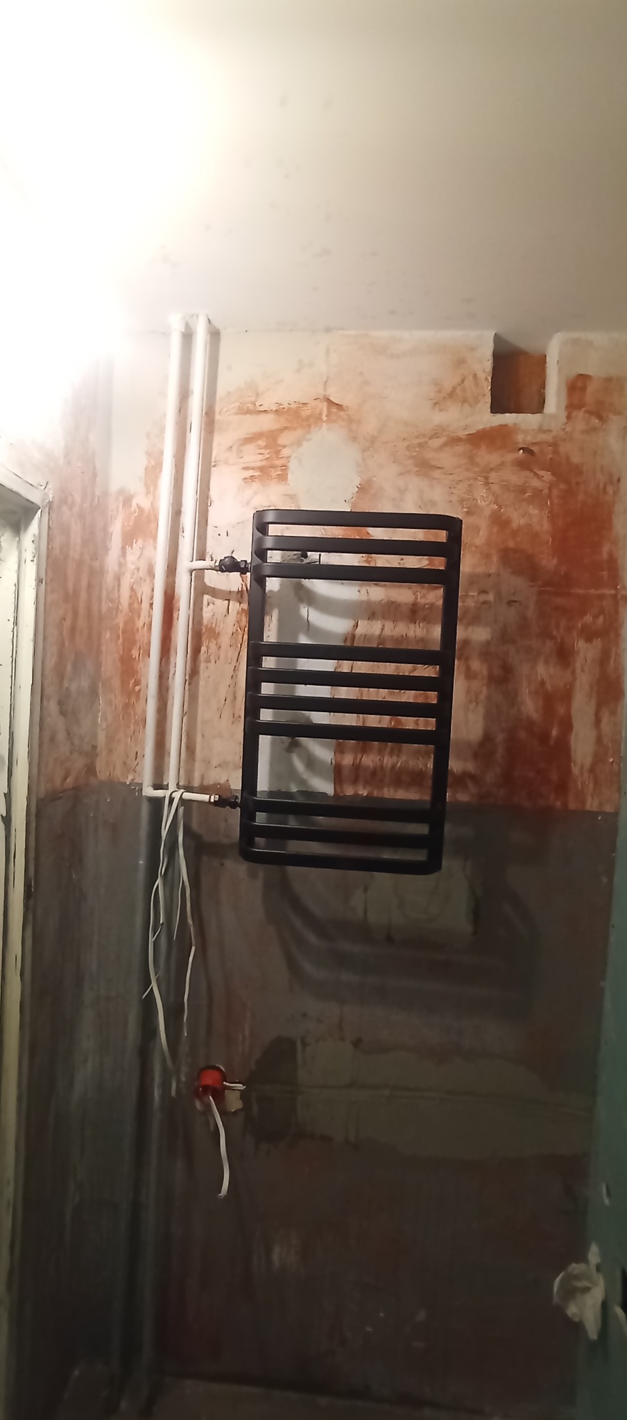 Ciemny grzejnik drabinkowy zamontowany na ścianie w trakcie remontu łazienki. Widoczne rury, surowe ściany z pozostałościami farby i instalacja elektryczna.