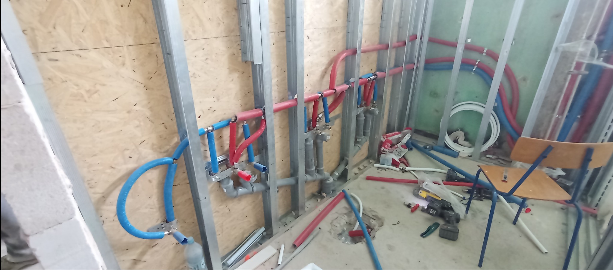 Instalacja wodno-kanalizacyjna w trakcie montażu: niebieskie i czerwone rury prowadzone wzdłuż metalowych profili, narzędzia na podłodze, widok z bliska.