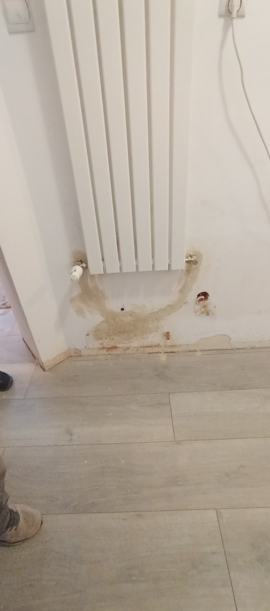Biały grzejnik panelowy na ścianie z widocznymi śladami po wycieku i uszkodzeniach tynku. Fragment podłogi z szarych paneli. Widoczne ślady po instalacji.