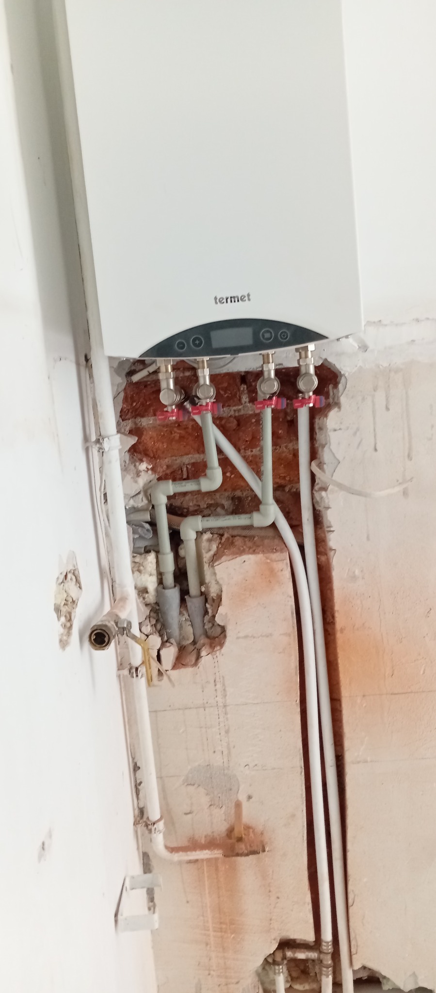 Biały piec gazowy Termet z podłączonymi rurami w surowym wnętrzu. Widoczne fragmenty ceglanej ściany i instalacji. Etap montażu w trakcie remontu.