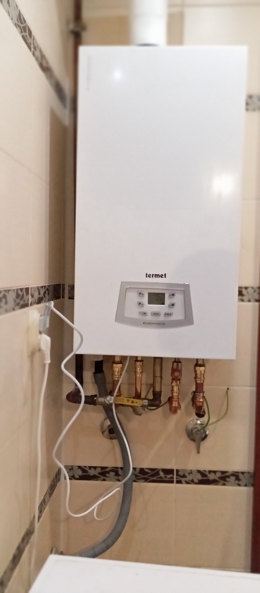 Biały, wiszący kondensacyjny piec gazowy Termet EcoCondens Gold Plus z widocznymi przyłączami hydraulicznymi na tle beżowych płytek w łazience.