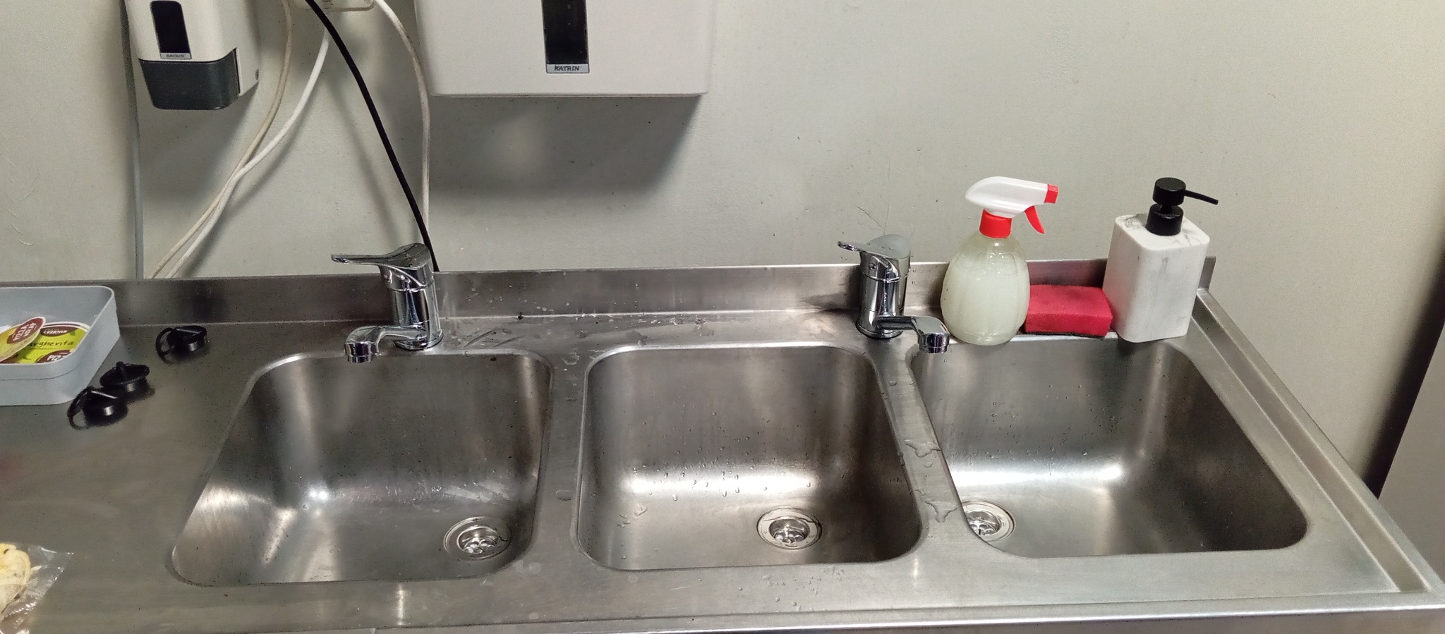 Trzykomorowy, stalowy zlew z bateriami, dozownikiem mydła i płynem do dezynfekcji. Widoczne ślady użytkowania i akcesoria czyszczące. Ujęcie z bliska, oświetlenie dzienne.