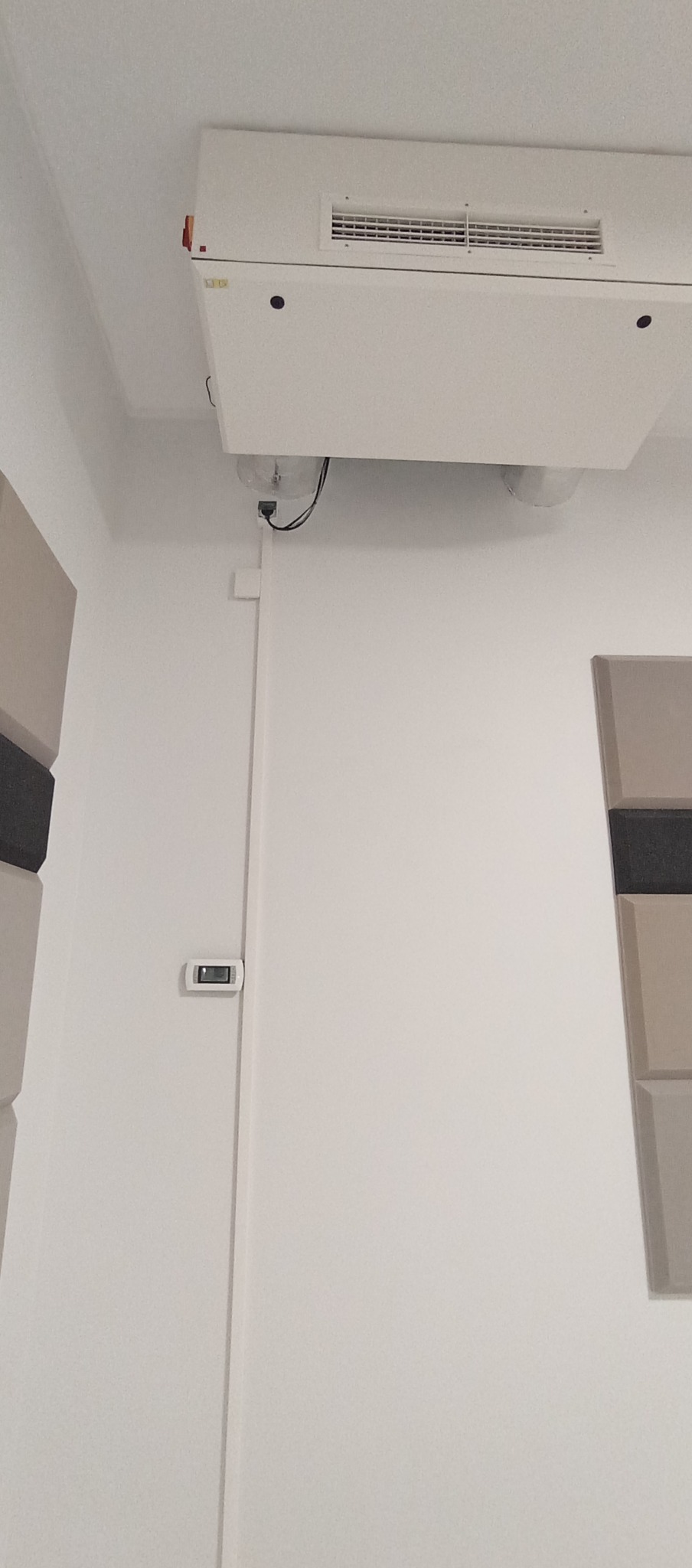 Biała jednostka klimatyzacyjna zamontowana pod sufitem, widoczny termostat na ścianie i panele akustyczne. Minimalistyczny design wnętrza.