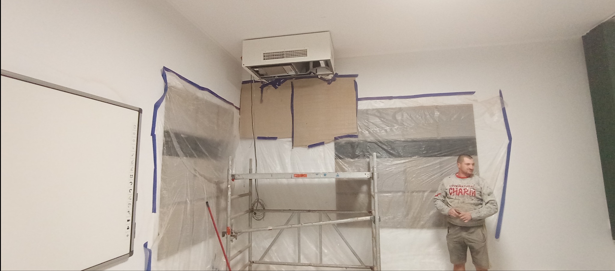 Przygotowania do remontu: zabezpieczone folią okna, rusztowanie, klimatyzator pod sufitem, mężczyzna w tle. Etap prac przed malowaniem ścian.