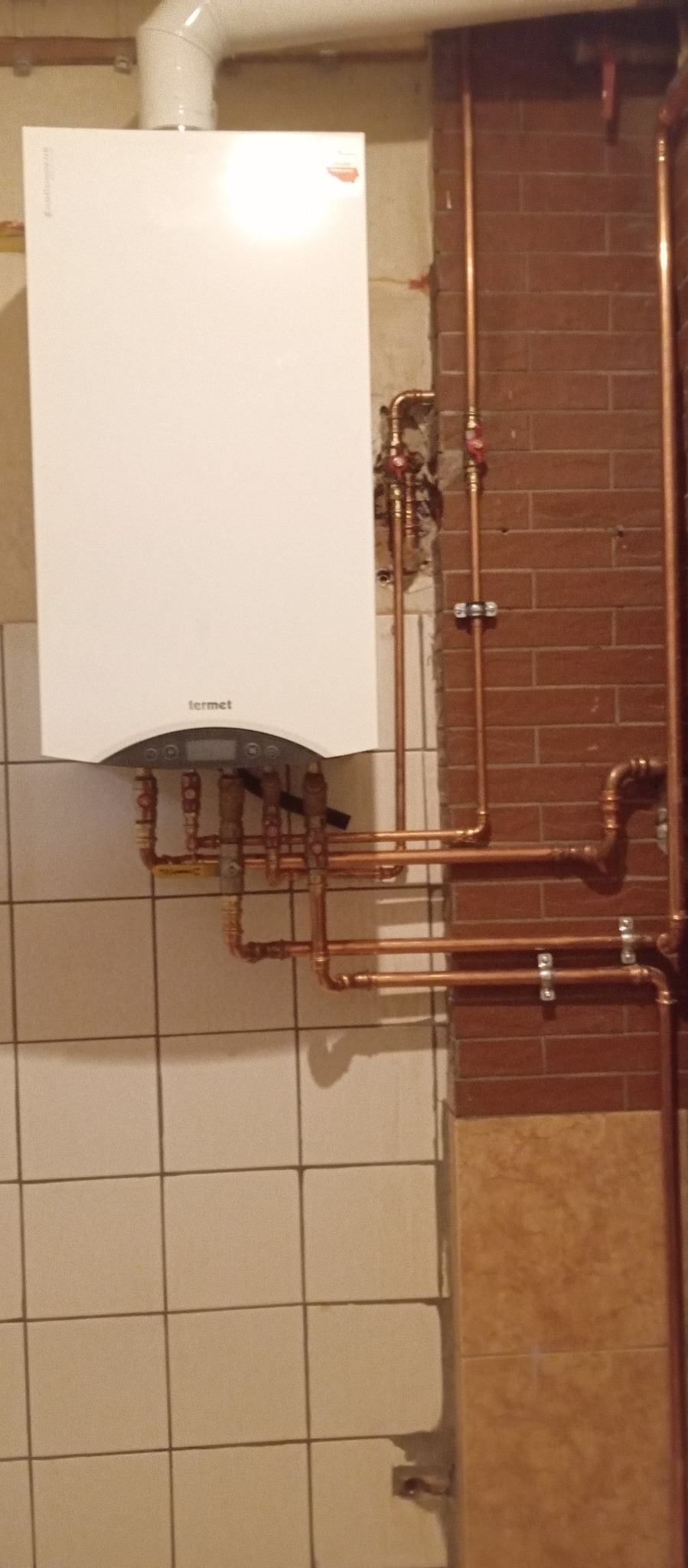 Biały piec gazowy Termet na tle ściany z cegły i płytek, z widocznymi miedzianymi rurami instalacji grzewczej. Ujęcie z bliska, prezentujące szczegóły podłączeń.