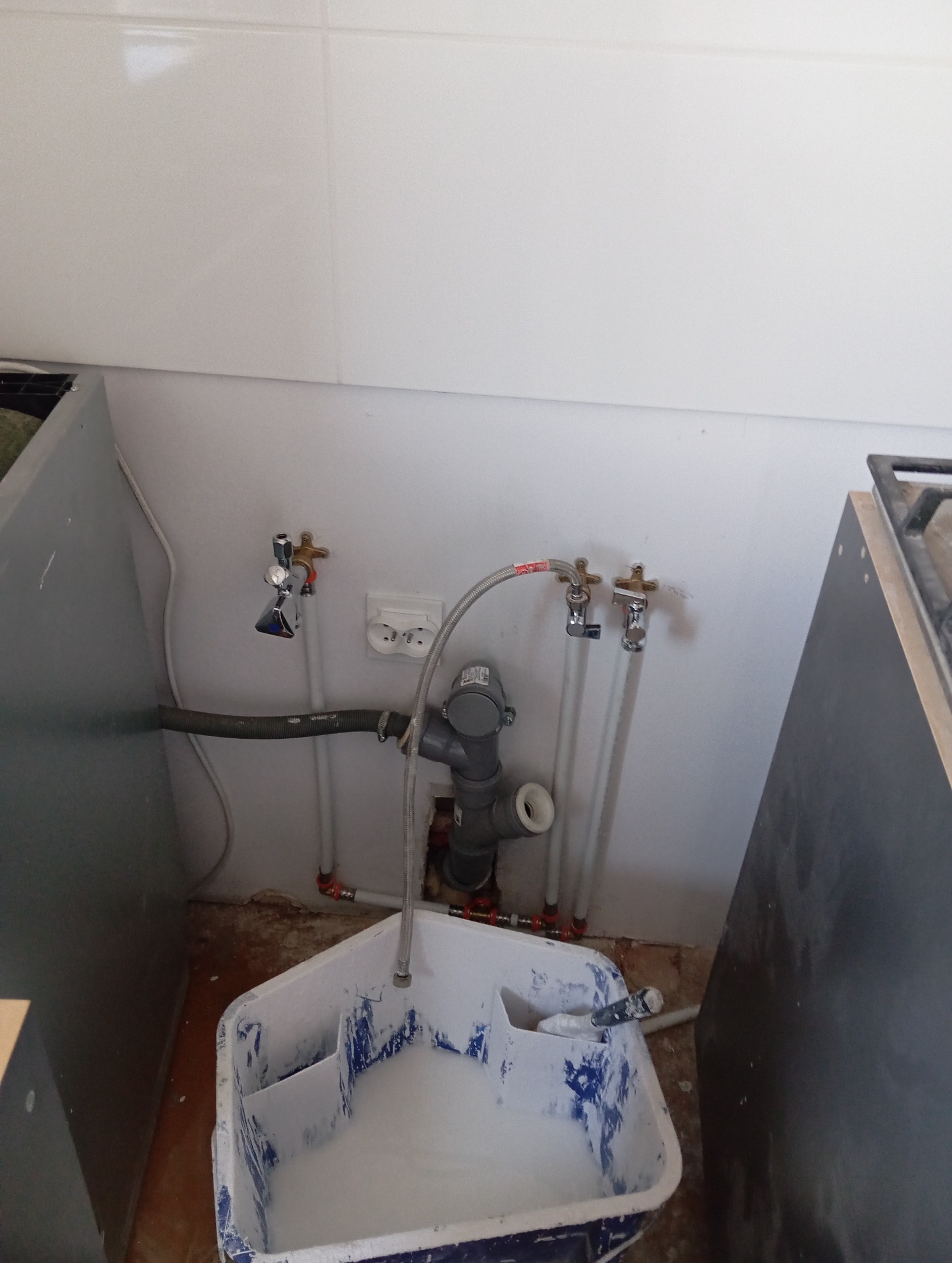 Fragment kuchni w trakcie prac hydraulicznych: widoczne rury, zawory, odpływ i wiadro z zaprawą. Ściany wyłożone białymi płytkami, podłoga w stanie surowym.