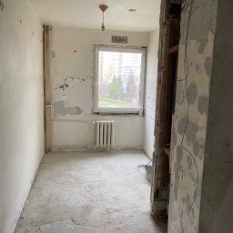 Qulfresh Krystian Kulikowski - Wnętrze mieszkania w trakcie demontażu, ściany częściowo obdarte z tapet, odsłonięta instalacja elektryczna, widoczny grzejnik pod oknem i fragment budynku za oknem.