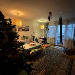 Qulfresh Krystian Kulikowski - Pokój z choinką, dywanem w orientalne wzory i psem leżącym na posłaniu przy balkonie, oświetlony trzema lampami z abażurami z wikliny.