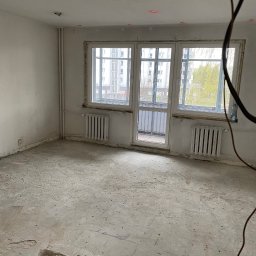 Qulfresh Krystian Kulikowski - Pusty pokój w mieszkaniu przed remontem z odsłoniętą podłogą, widoczne ślady po instalacjach na suficie i czerwone punkty laserowe, okna z widokiem na blok.