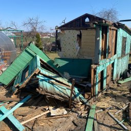 Qulfresh Krystian Kulikowski - Zniszczony, drewniany budynek z odpadającym zielono-niebieskim malowaniem, odsłoniętą tapetą w kwiaty, gruzem i elementami konstrukcyjnymi porozrzucanymi wokół, na tle błękitnego nieba.