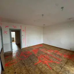 Pokój przed remontem z zaznaczonymi miejscami do wyburzenia czerwoną farbą na ścianach i podłodze. Widoczne stare podłogi i drzwi.