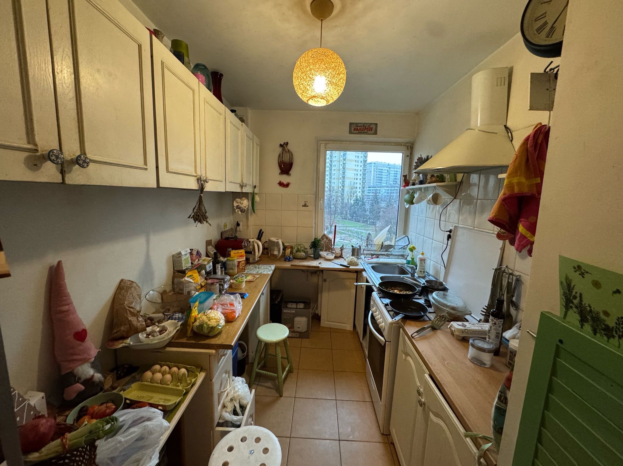 Widok na zagraconą kuchnię z białymi szafkami, brązowymi płytkami podłogowymi i widokiem na osiedle za oknem.