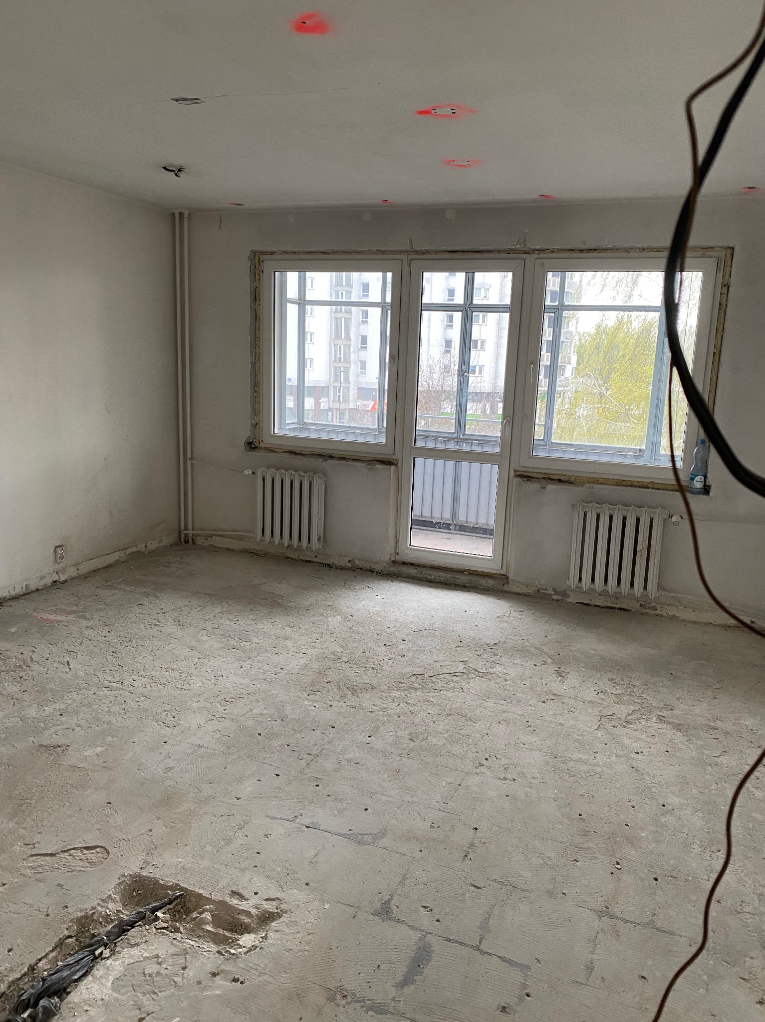 Pusty pokój w mieszkaniu przed remontem z odsłoniętą podłogą, widoczne ślady po instalacjach na suficie i czerwone punkty laserowe, okna z widokiem na blok.