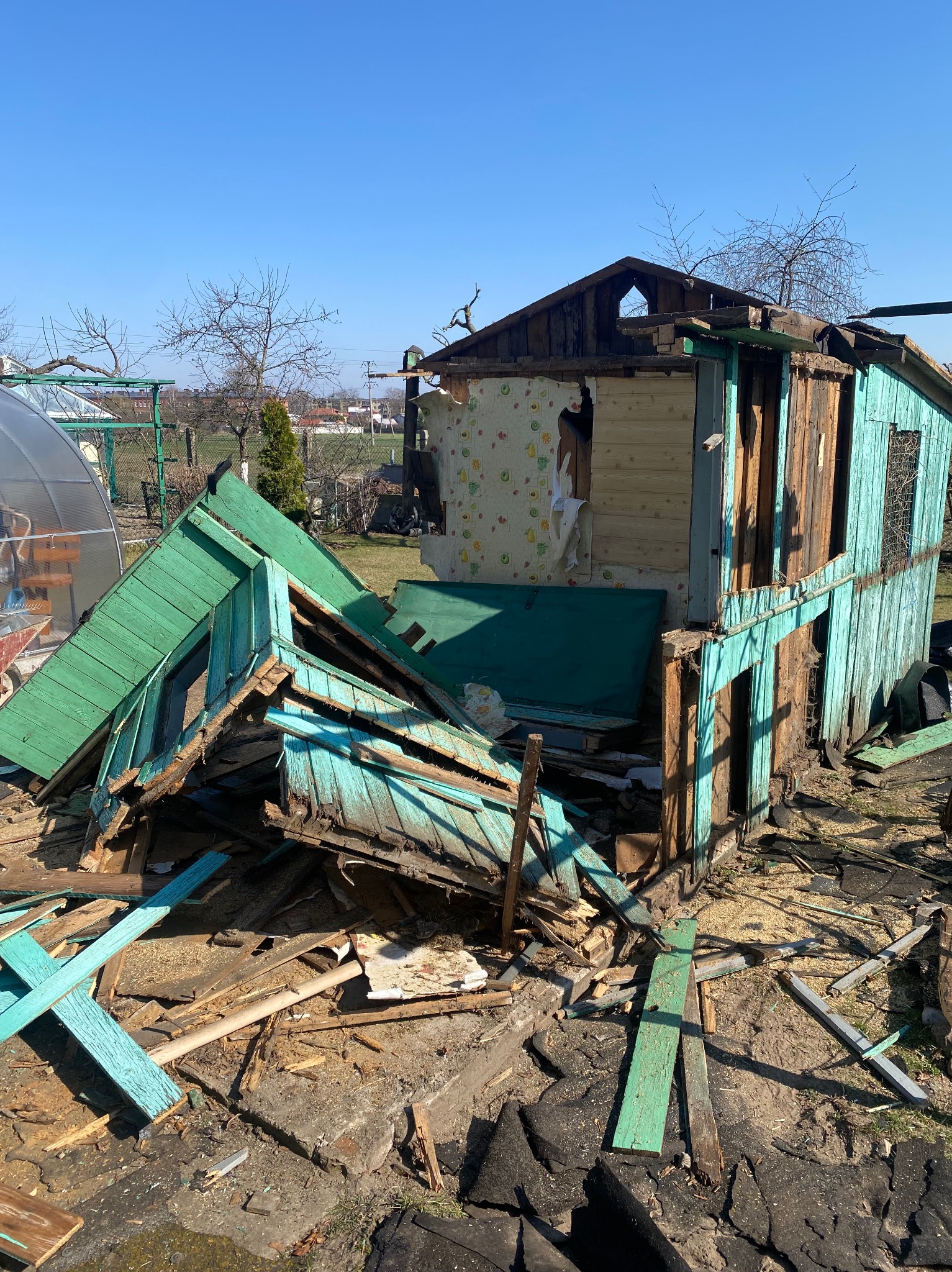 Zniszczony, drewniany budynek z odpadającym zielono-niebieskim malowaniem, odsłoniętą tapetą w kwiaty, gruzem i elementami konstrukcyjnymi porozrzucanymi wokół, na tle błękitnego nieba.