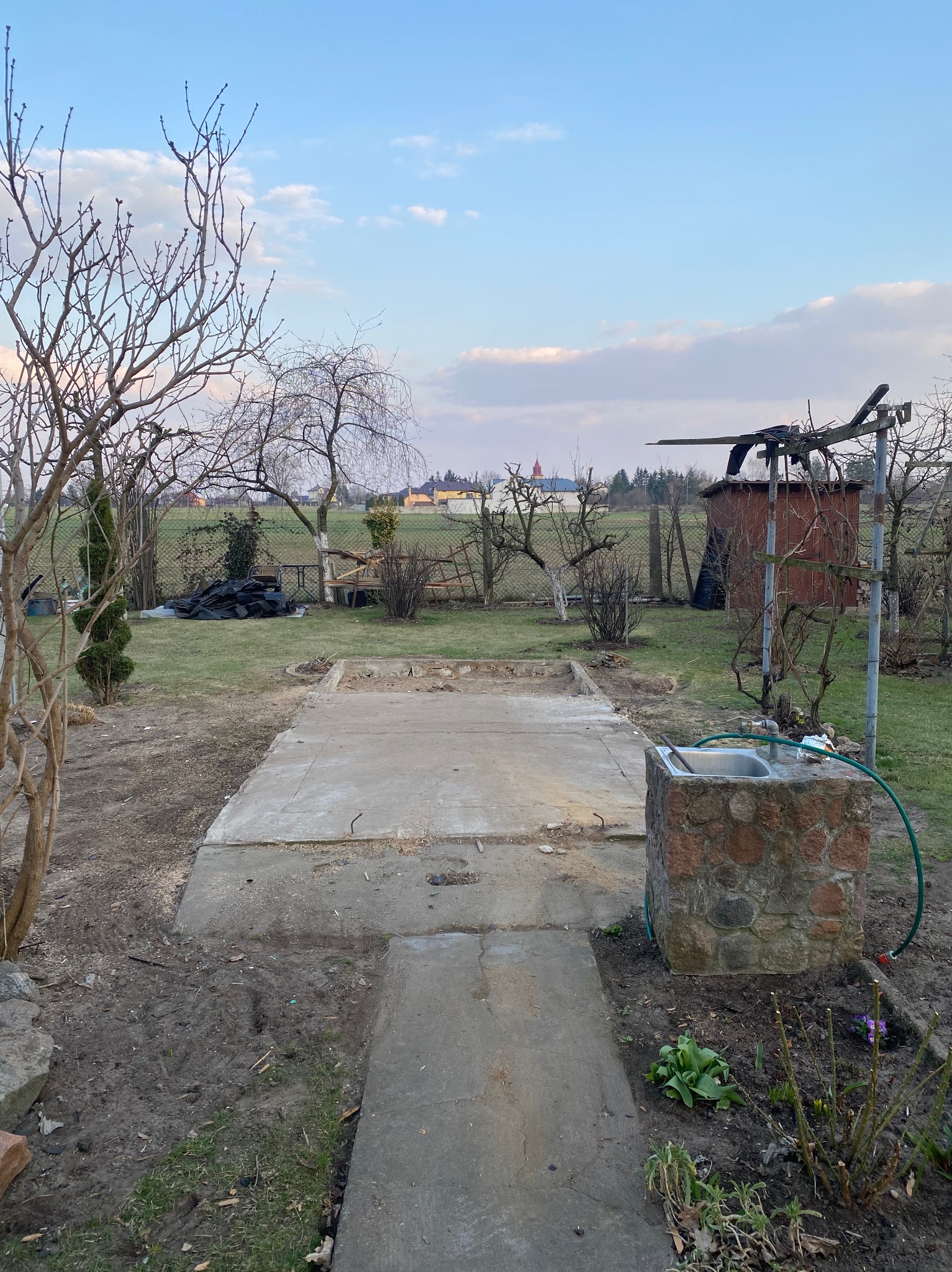 Betonowy fundament w ogrodzie z widocznym zlewem z kamienia i zaroślami, w tle domy i kościół pod błękitnym niebem.