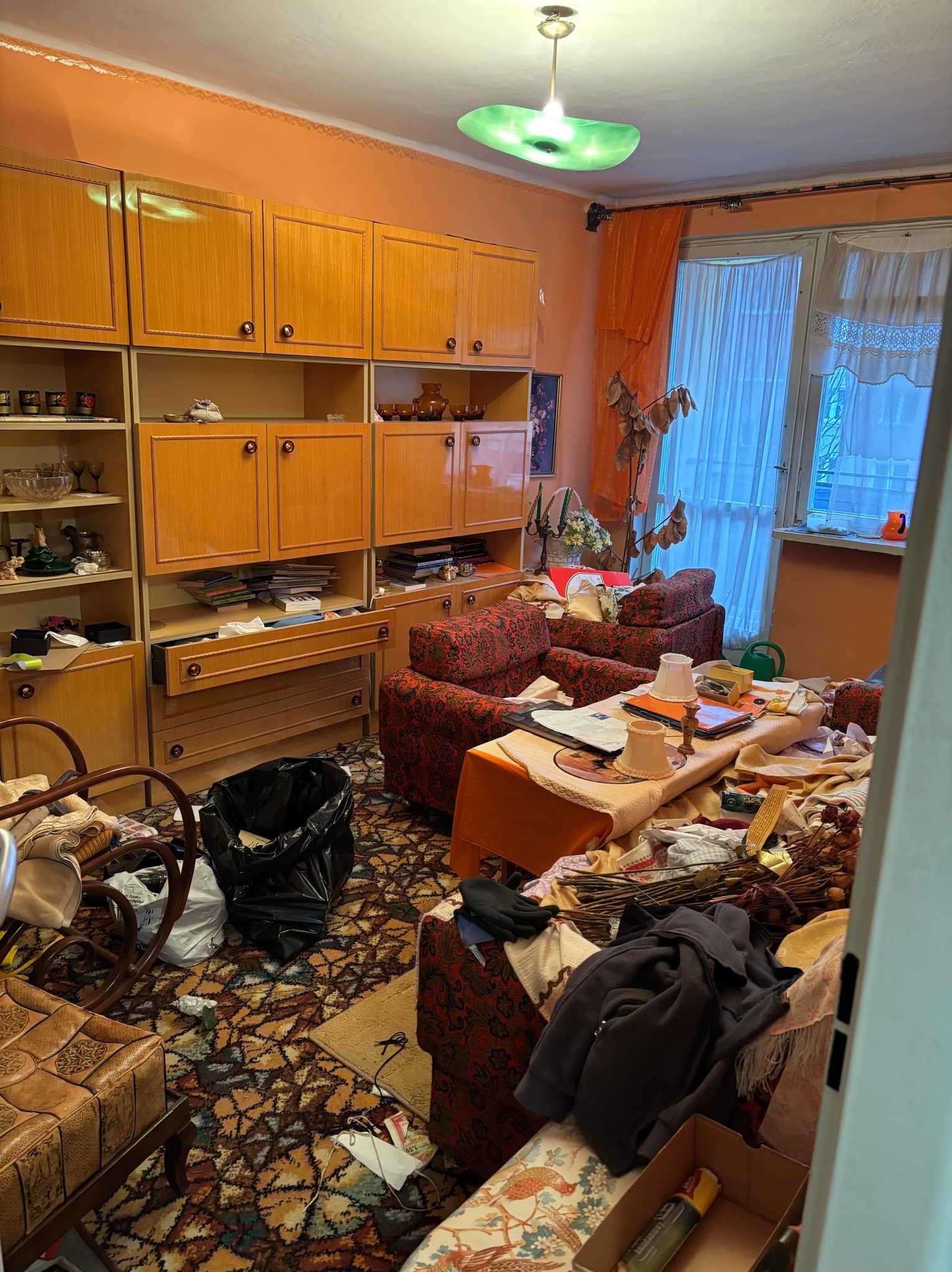 Zaniedbane wnętrze pokoju z meblościanką, wzorzystą tapetą, sofą w kwiaty, stolikiem zastawionym przedmiotami i czarnym workiem na śmieci; widok zza uchylonych drzwi.