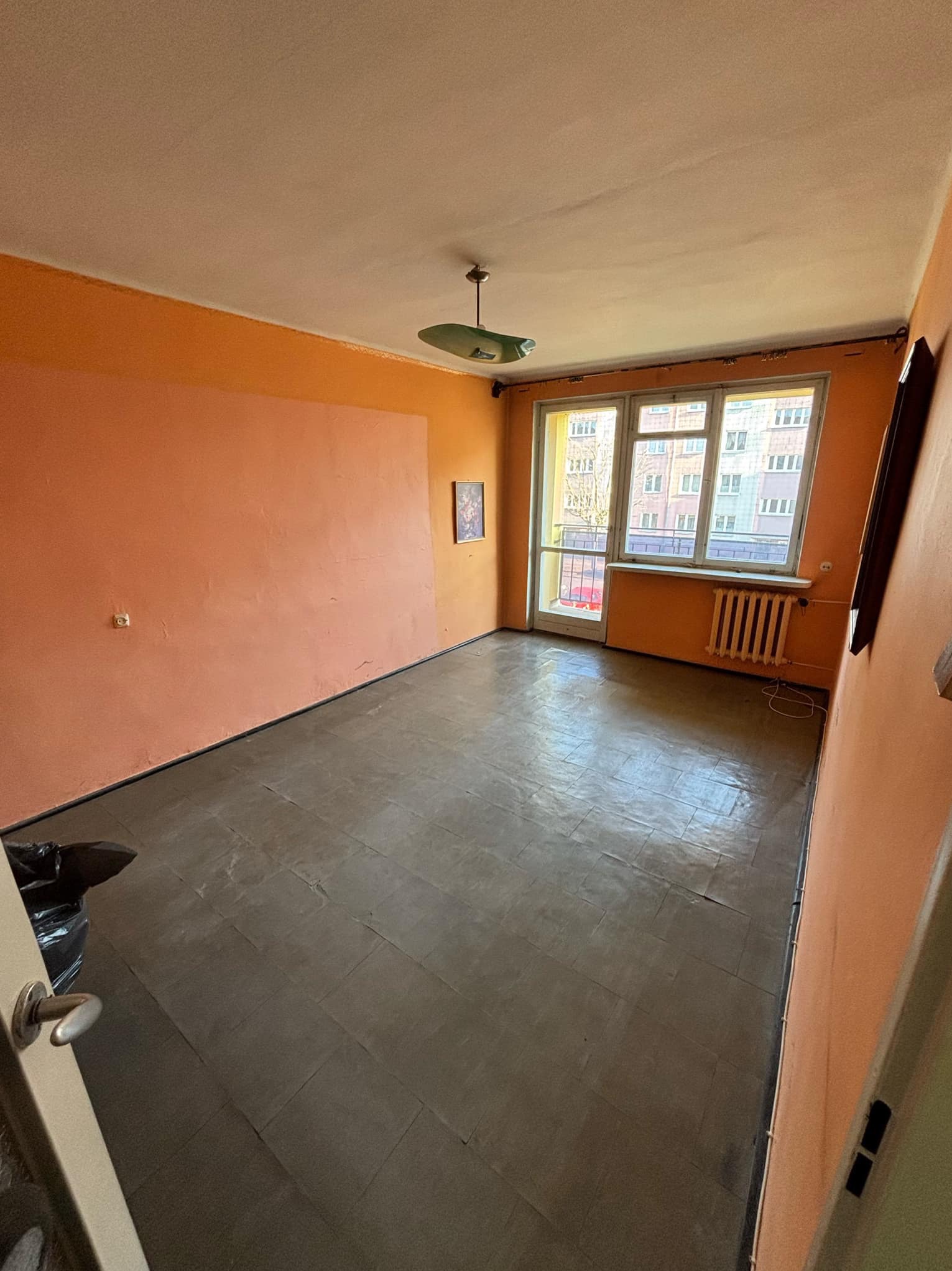 Pusty pokój z pomarańczowymi ścianami, brązową podłogą i oknem balkonowym, widoczny czarny worek na śmieci, przygotowany do opróżnienia.