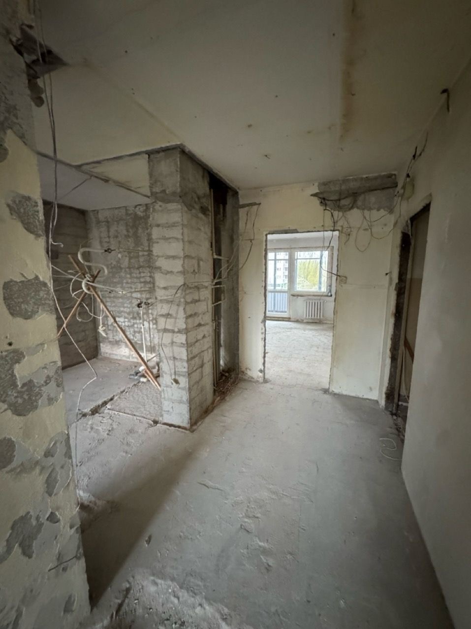 Wnętrze po demontażu ścian działowych, widoczne odsłonięte kable i resztki tynku. Świeży beton na podłodze, okno w tle. Ujęcie z perspektywy wnętrza.