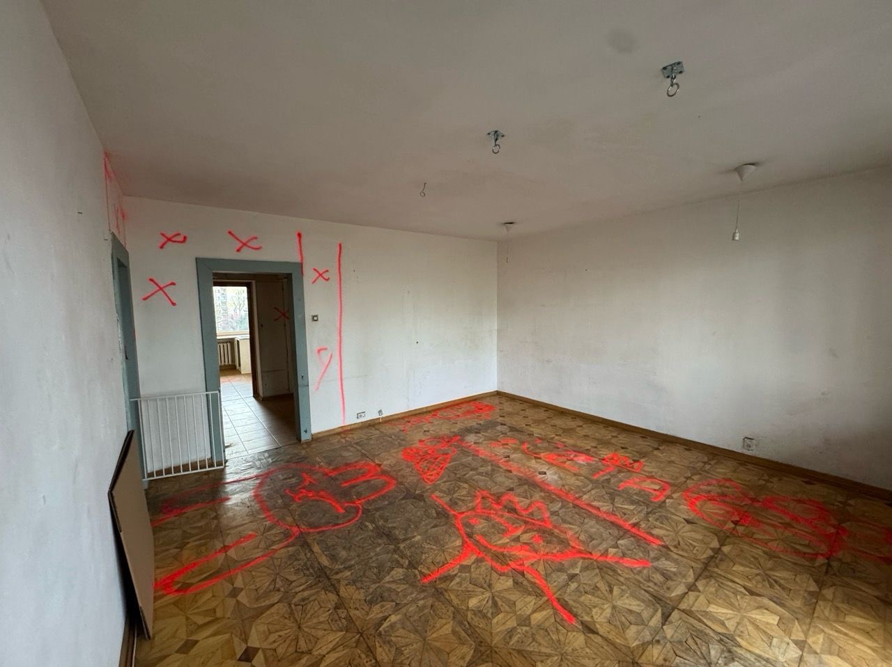 Pokój przed remontem z zaznaczonymi miejscami do wyburzenia czerwoną farbą na ścianach i podłodze. Widoczne stare podłogi i drzwi.
