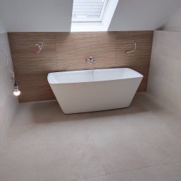 REMONTY A-Z Andrzej Kiewra - Nowoczesna, wolnostojąca wanna na tle ściany z drewnopodobnych paneli w jasnej łazience z oknem dachowym. Widoczne niedokończone instalacje elektryczne.