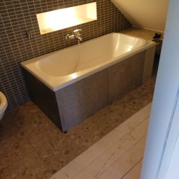 REMONTY A-Z Andrzej Kiewra - Biała wanna obudowana szarymi płytami w łazience na poddaszu, ściana wyłożona mozaiką, wnęka z oświetleniem. Widoczne płytki podłogowe i fragment białych desek.