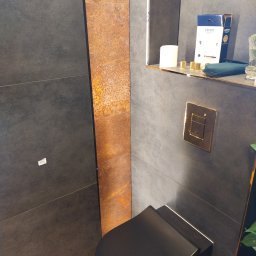 REMONTY A-Z Andrzej Kiewra - Nowoczesna łazienka z czarną miską WC, złotym przyciskiem spłukującym i płytkami w odcieniach szarości oraz rdzawego brązu. Widoczna półka z akcesoriami.