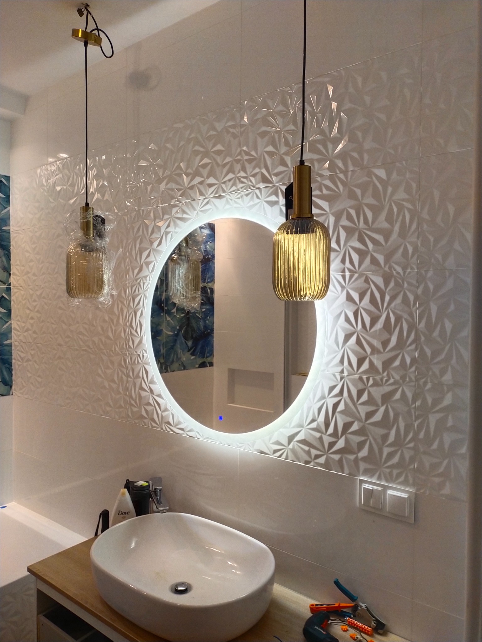 Nowoczesna łazienka z umywalką nablatową, okrągłym lustrem z podświetleniem LED i dekoracyjnymi, trójwymiarowymi płytkami ściennymi. Wiszące lampy w złotym kolorze.