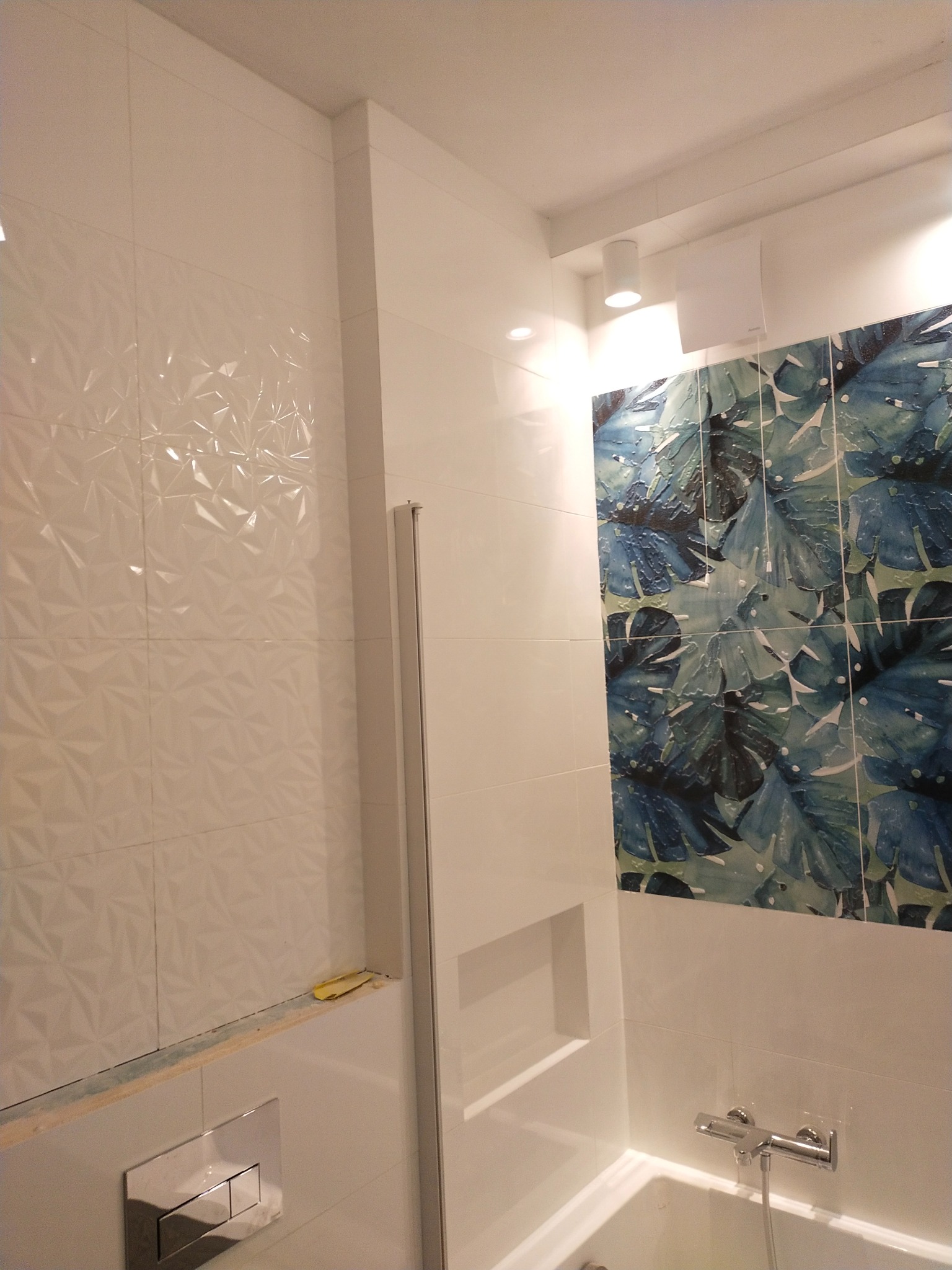 Biała łazienka z akcentem w postaci płytek z motywem liści. Widoczne geometryczne płytki 3D i wnęka w ścianie. Minimalistyczny design i nowoczesne wykończenie.