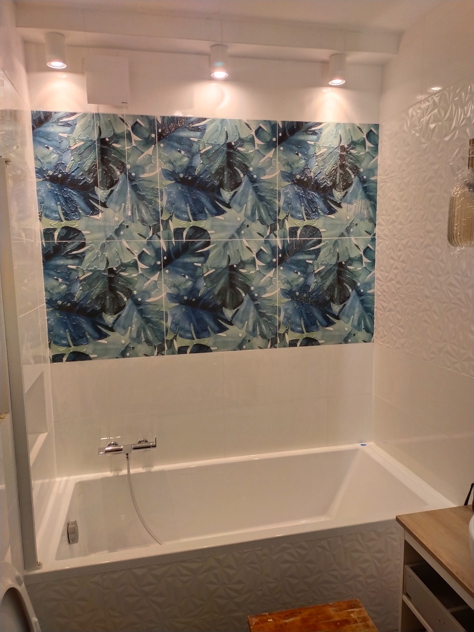 Biała wanna z baterią, otoczona płytkami: gładkie białe, wzorzyste z motywem liści i trójwymiarowe białe. Oświetlenie punktowe na suficie.