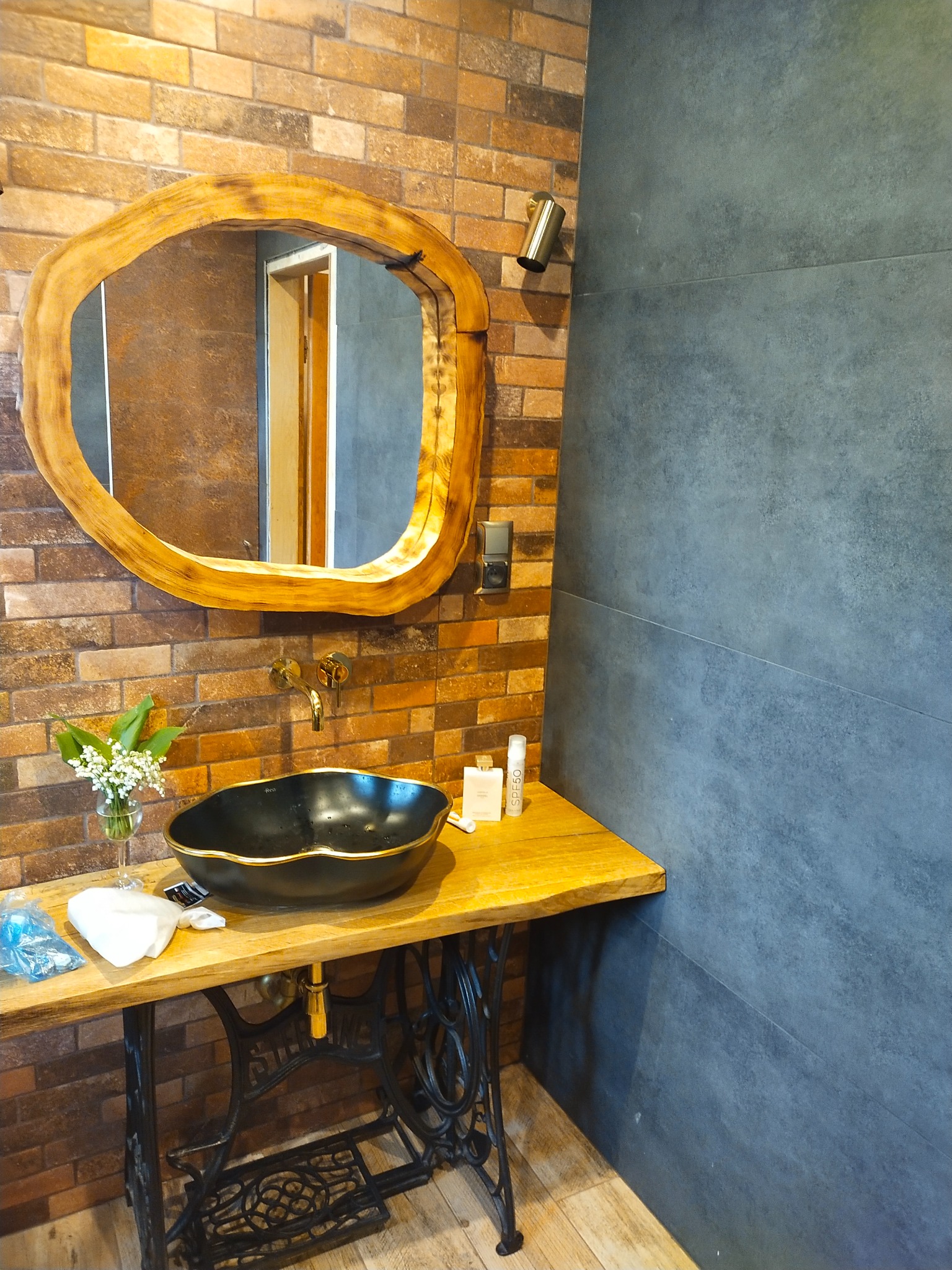 Rustykalna łazienka z umywalką w stylu retro na podstawie maszyny do szycia, lustrem w drewnianej ramie i ścianą z cegły oraz szarych płytek.