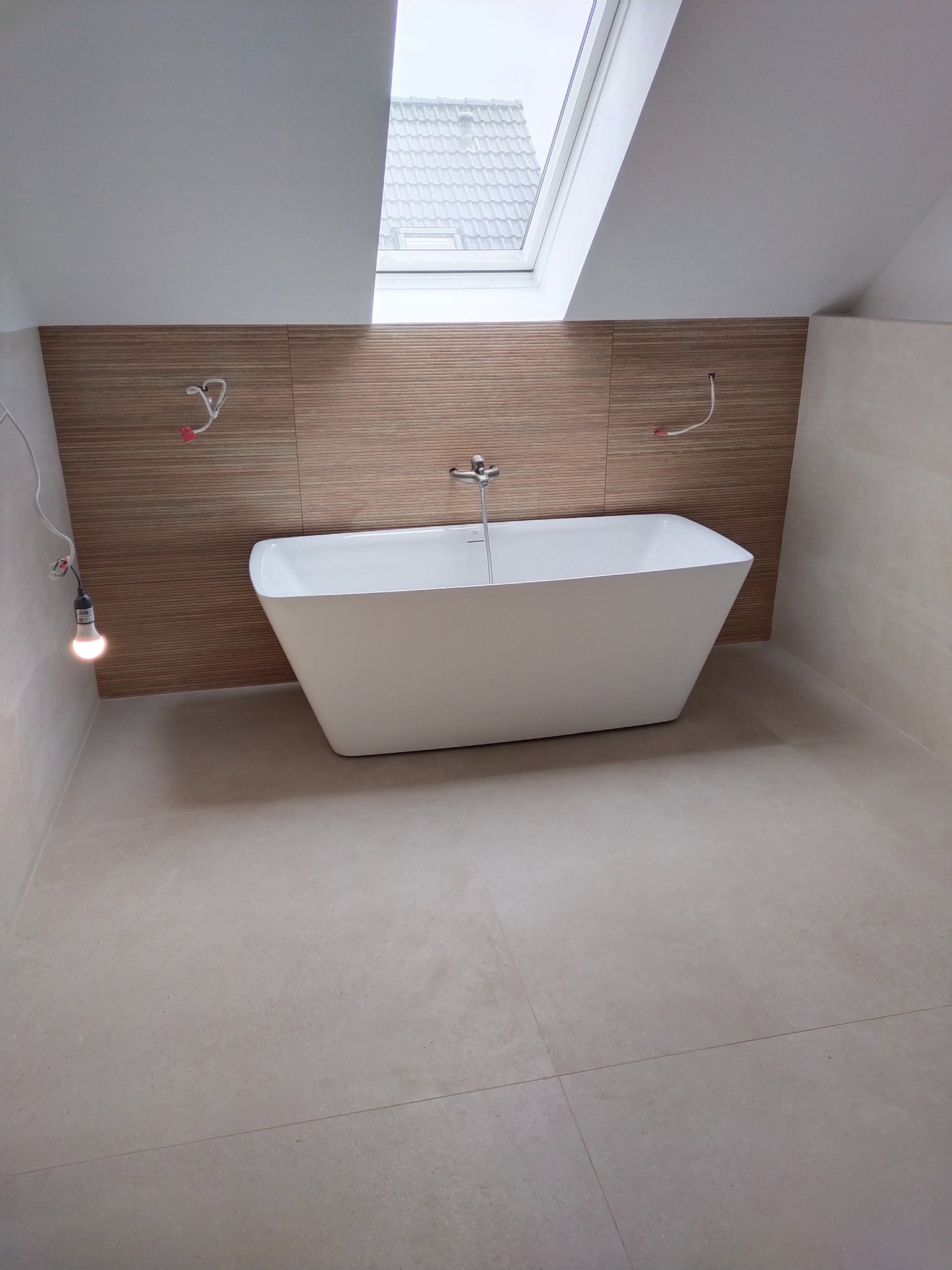 Nowoczesna, wolnostojąca wanna na tle ściany z drewnopodobnych paneli w jasnej łazience z oknem dachowym. Widoczne niedokończone instalacje elektryczne.