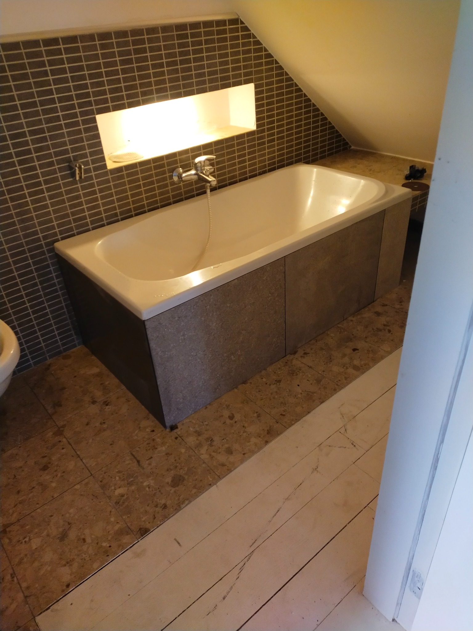 Biała wanna obudowana szarymi płytami w łazience na poddaszu, ściana wyłożona mozaiką, wnęka z oświetleniem. Widoczne płytki podłogowe i fragment białych desek.