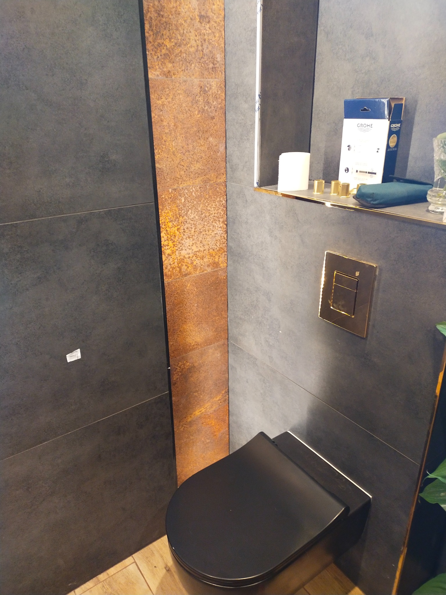 Nowoczesna łazienka z czarną miską WC, złotym przyciskiem spłukującym i płytkami w odcieniach szarości oraz rdzawego brązu. Widoczna półka z akcesoriami.