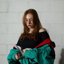 Kub1k - Młoda kobieta z rudymi włosami, ubrana w czarny top z odkrytymi ramionami, zapinająca zielono-czerwono-białą kurtkę na tle białej, ceglanej ściany.